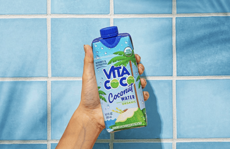 Eine Hand präsentiert eine Vita Coco-Kokoswasserpackung vor einem blau gekachelten Hintergrund, um die Attraktivität des Produkts in der wettbewerbsintensiven Welt des CPG-Getränkemarketings hervorzuheben.
